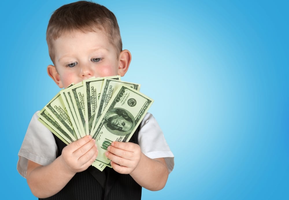 a toddler wearing a white shirt and black vest holding a handful of $100 bills spread out like a fan