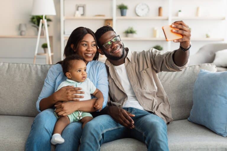 Portrait of cute African American dad and mom taking selfie with their kid at home, cheerful black family having good time together, taking photos on smartphone camera, sitting on couch in living room